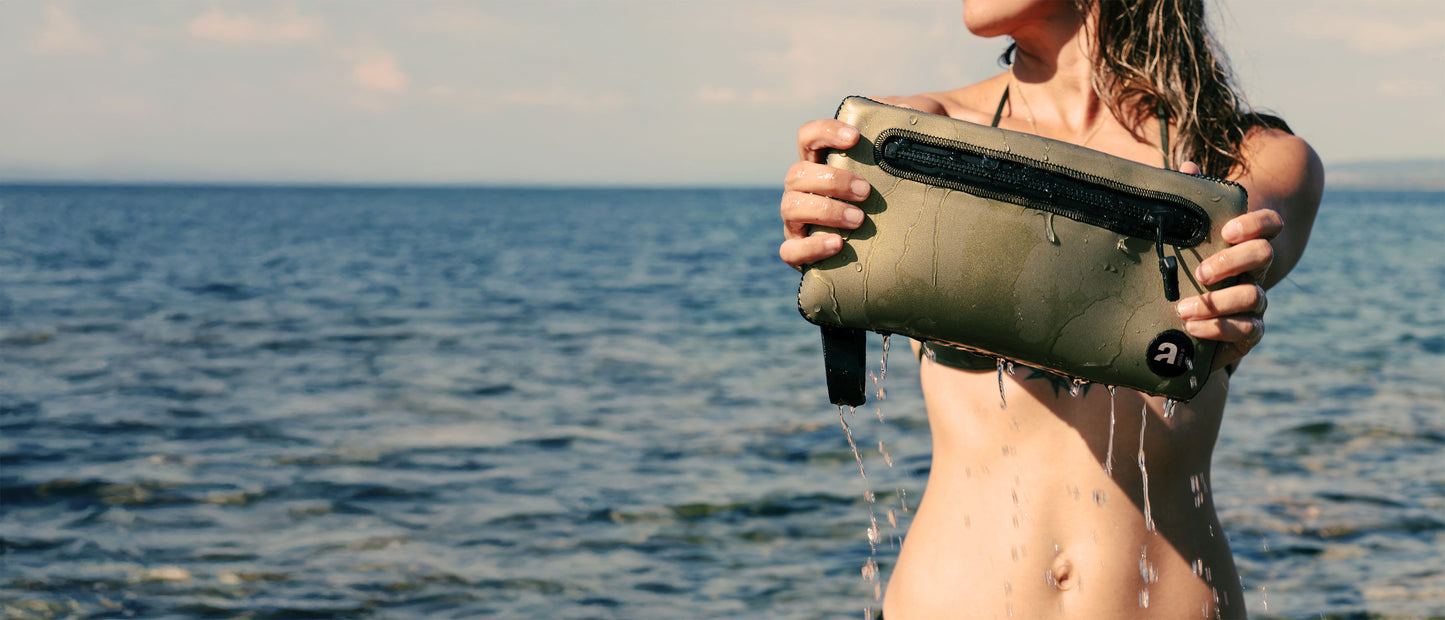 Ragazza in riva al mare tiene in mano la pochette Lise in neoprene glideskin oro.