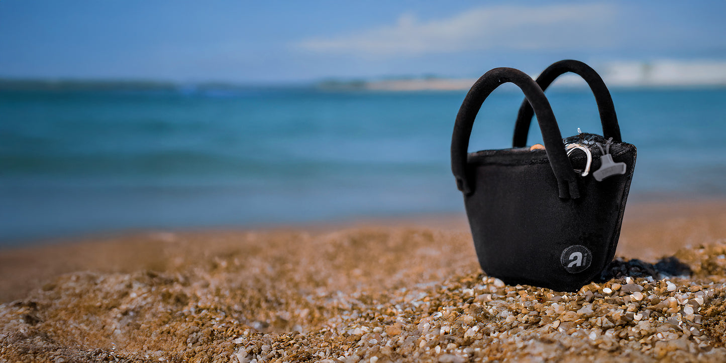 Borsa a secchiello Iris in neoprene glideskin nero di Atelis, appoggiata sulla sabbia in riva al mare.